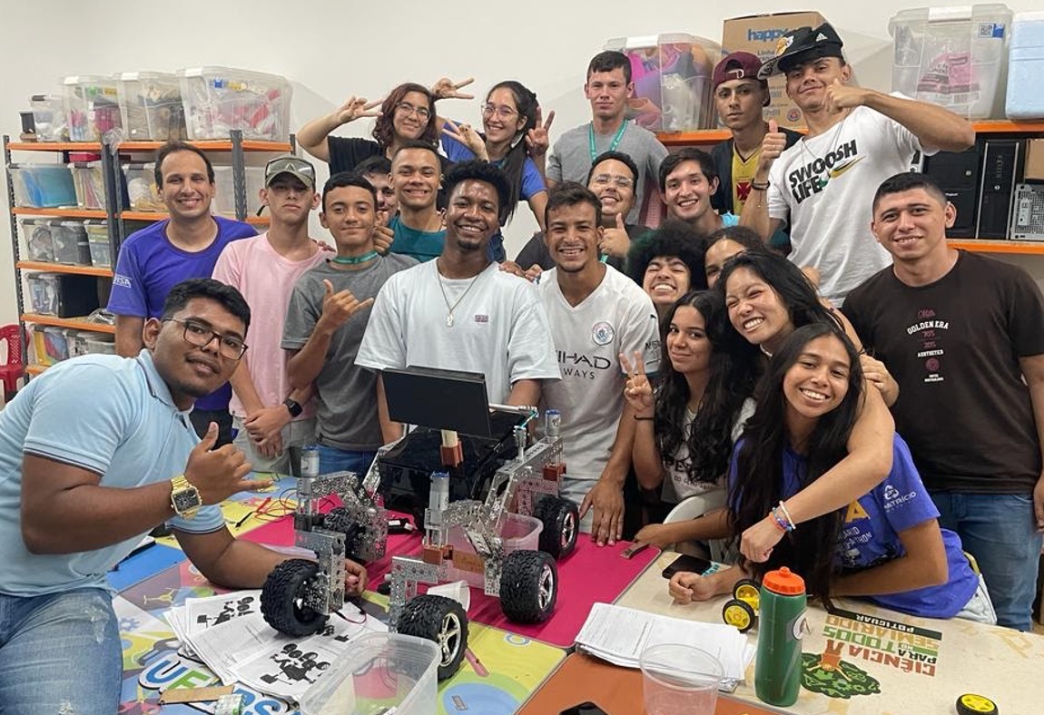 Manuel Morales among the 19 high school students that worked on a Open Source Rover on the table with 4 small wheels and a rectangular screen atop of it