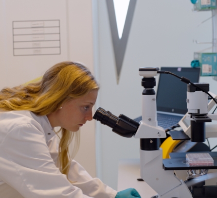 Meredith Arterburn in a white lab coat crouching over a table with a microscope and looking through it