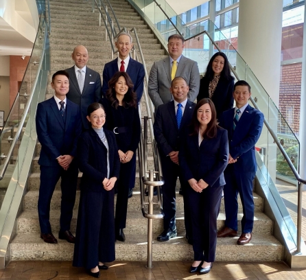 Christine Pilcavage standing at the bottom of the stairs to the left in a grey turtleneck and a suit with one hand on top of the other. Chris is standing with nine other Japanese American leaders in suits