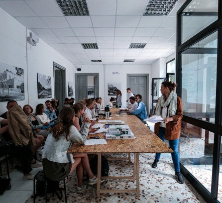 Two seated rows of MIT students sitting to the left of a long rectangle wood table while two seated at the front and another two people in the front and back standing to the right of the table in a room with floor-to-ceiling glass windows to the right with sun shining in