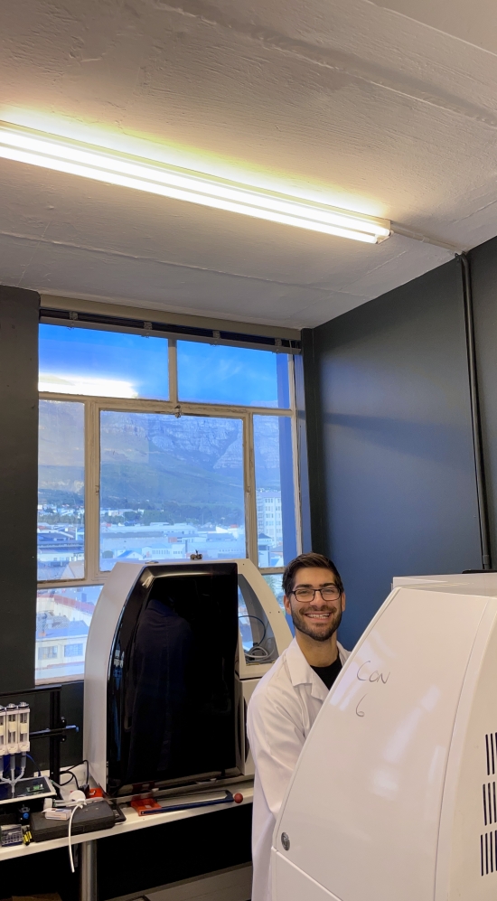 Branden Spitzer in a lab coat working behind a bioreactor at Mzansi Meat Co. with a birds-eye view of Table Mountain in Cape Town, South Africa