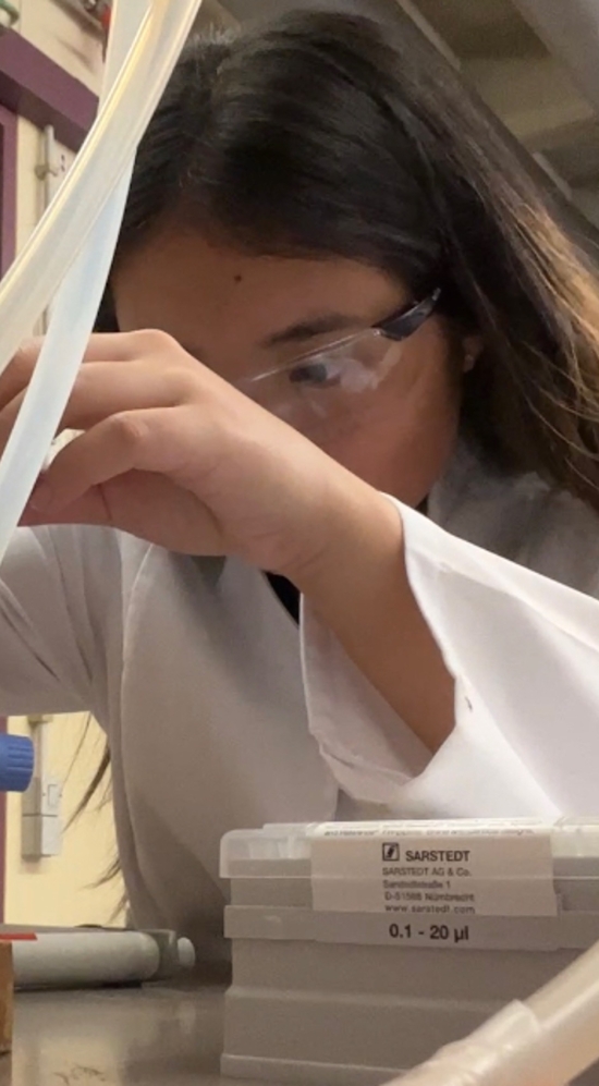 Jessie Liu wearing a white lab coat and goggles while handling a white long pippette-like equipment on a table with tubes and a fire extinguisher hung against the wall next to her work table