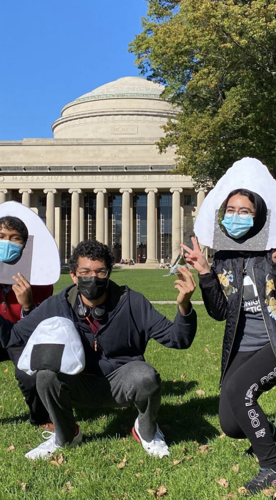 Marwa AlAlawi wearing a onigiri photobooth prop holding two peace signs with both hands crouching down halfway to the right with two other MIT students who's also crouching down on the grass, one wearing a onigiri photobooth prop and another with glasses holding an onigiri rice ball in one hand and rested on his thigh, an onigiri pillow in front of Killian Court at MIT