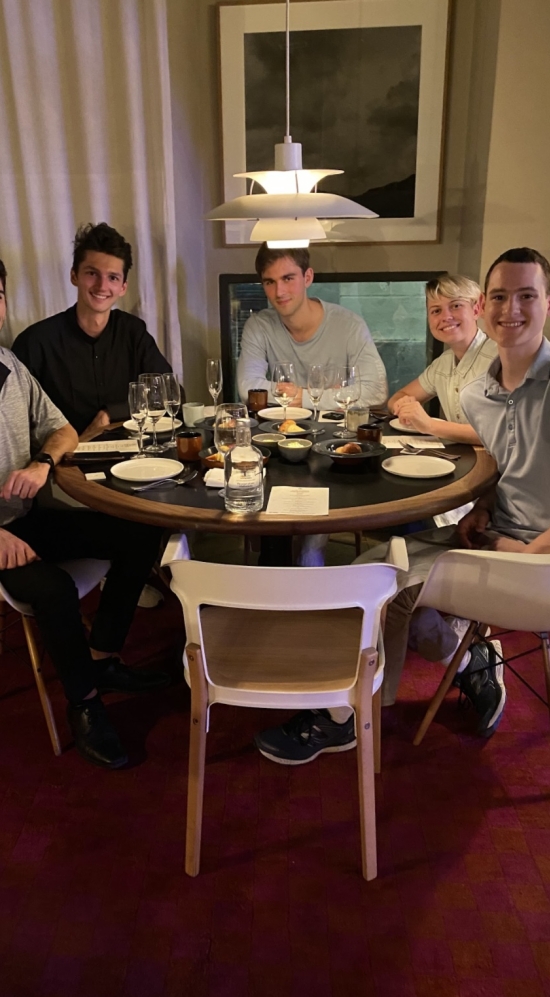 Graham Guite and MIT peers sitting around a round table for dinner in Barcelona, Spain in a room with red carpet and a low-hanging lamp 