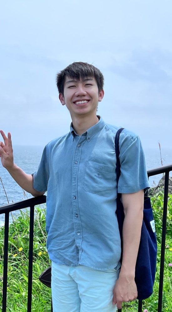 Michael Tan holding up a peace sign as he stands at the fence overlooking the sea and a grass-covered cliff