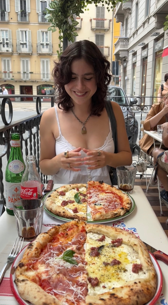 Layla smiling down at two pans of a two-halves pizza combination at a table on a patio in Milan, Italy