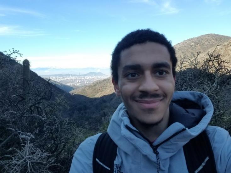 A selfie of Kinan Martin hiking at Las Aguas Ramones with dry branches behind him and in a distant a city of Santiago, Chile